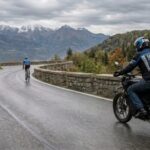 A motorcycle on the road behind the bicyclist in a rainy mountain terrain.