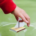 A child's hand placing a paper boat in the pond.