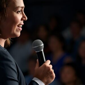 A close-up, clean, professional photograph of a person mid-speech holding a microphone, looking calm, grounded, and confident with relaxed shoulders. Audience is visible, though blurred.