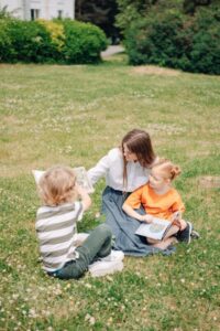 A mother reads storybooks to her children while sitting on lush green grass outdoors, fostering learning.
