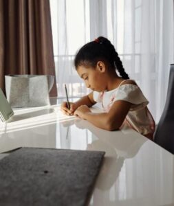 A young girl concentrates on her homework at home near a window.