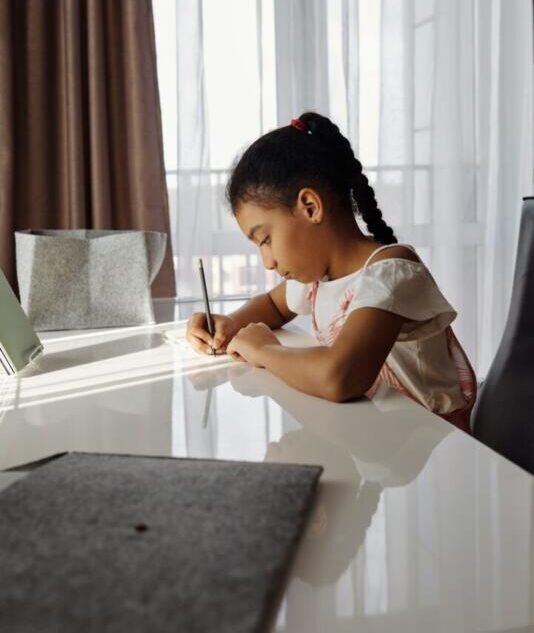 A young girl concentrates on her homework at home near a window.