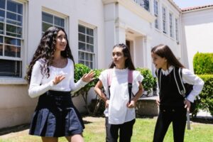 Three teenage girls in uniforms having a lively discussion outside their school on a sunny day.