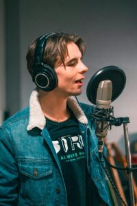 Voice actor in blue denim jacket standing at a microphone with a pop screen.