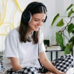 Young woman with headphones working on a laptop in a cozy bedroom setting.