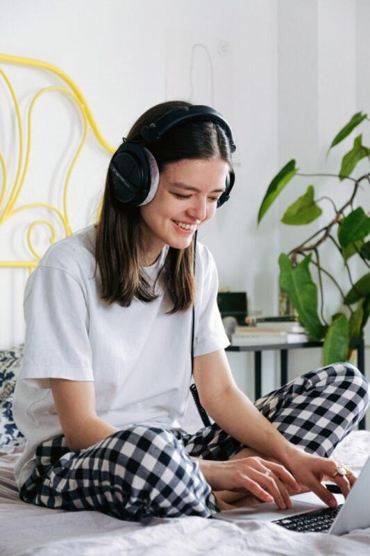 Young woman with headphones working on a laptop in a cozy bedroom setting.