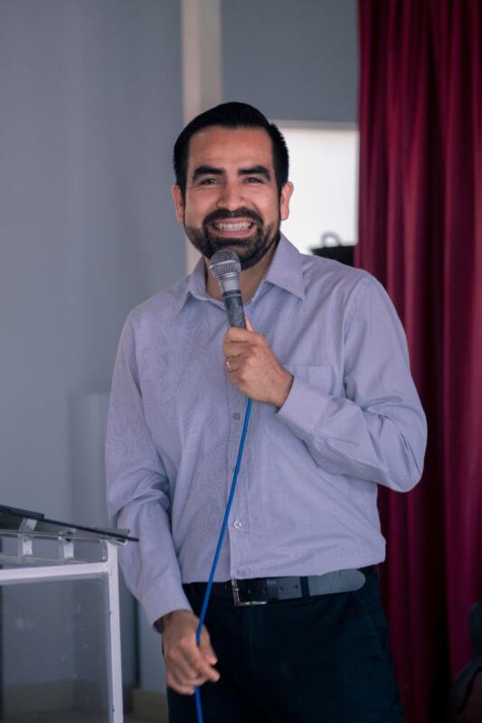 An energetic older adult speaking confidently into a microphone or addressing an engaged audience in a brightly lit room.