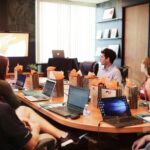 A diverse professional speaking confidently at a boardroom table with colleagues actively listening.