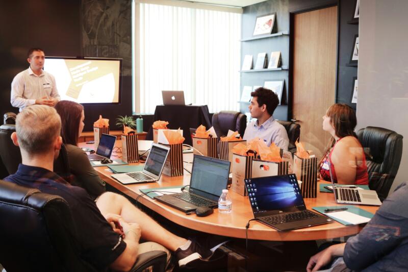 A diverse professional speaking confidently at a boardroom table with colleagues actively listening.