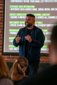 A person speaking comfortably with relaxed, confident posture in a warm indoor or office setting. Carlos Gil presenting insights on integrating SMS marketing for enhanced communication at Inbox Expo 2024.