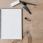 Flat lay of school supplies including notebook, compass, eraser, and protractor on wooden desk.