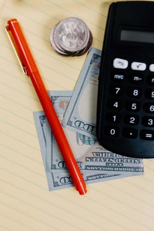 Close-up of a pen, calculator, and cash on a notepad.