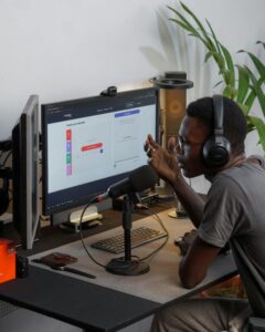 Man in dark crew neck t-shirt wearing black headphones talking into microphone sitting in a home workspace.