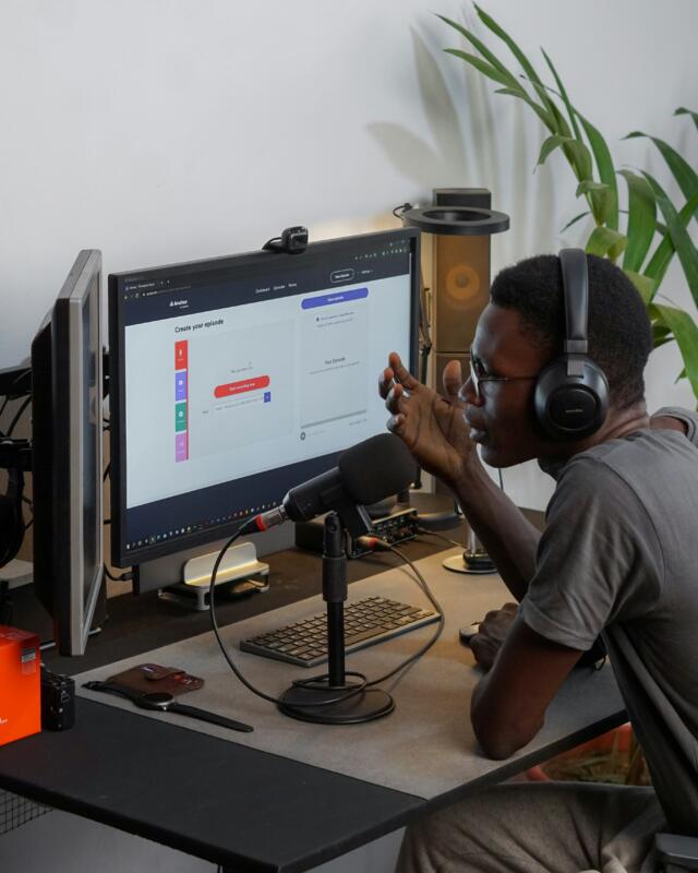Man in dark crew neck t-shirt wearing black headphones talking into microphone sitting in a home workspace.