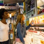 A pair of female friends in front of a taco truck looking at the menu.