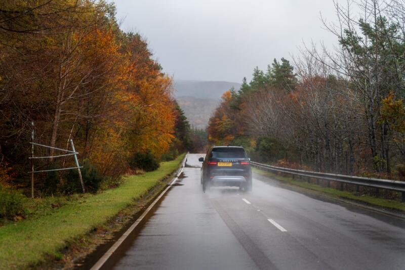 Black car driving down a wet country road during a daytime rainstorm.